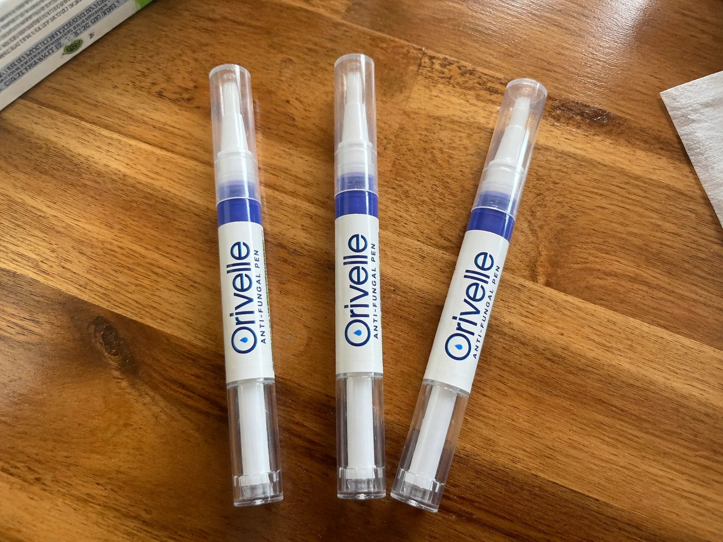 Phone snapshot of three Orivelle applicator pens laid out in a loose row on a warm wooden kitchen counter, showing the three-pack most patients order.