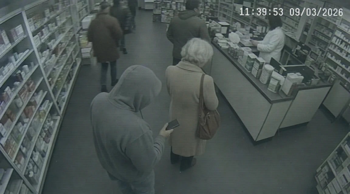 Full-frame low-resolution CCTV still in a small neighborhood pharmacy, high angle from a ceiling camera, desaturated cool grey-green, burned-in white timestamp 11:39:53 in the upper right; a silver-haired woman in a knee-length beige wool coat stands third in line holding a brown leather tote with both hands, and a man in a grey hooded sweatshirt stands roughly three feet behind her holding a small dark deck-of-cards-size rectangle at hip level angled toward her tote