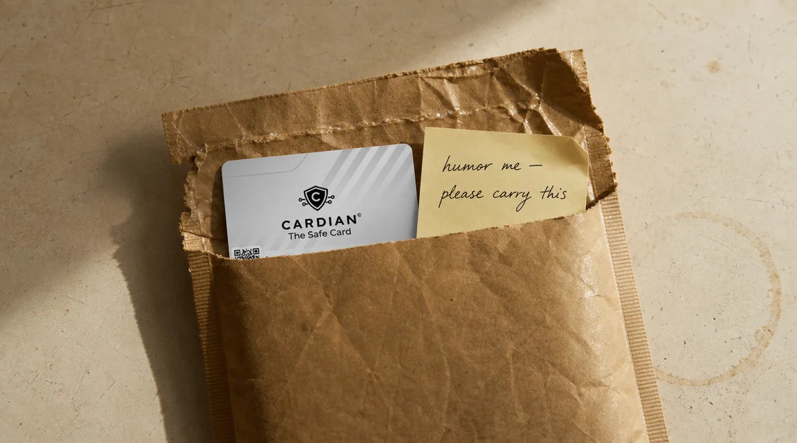 Editorial documentary still-life: a brown padded mailer torn open on a warm cream kitchen counter, a real Cardian card visible at the top of the envelope (white + grey diagonal stripes + black shield + CARDIAN wordmark + QR code), beside it a canary-yellow Post-it note with a handwritten message reading 'humor me — please carry this'. Late afternoon natural light, single coffee-ring stain on the counter. Atlantic editorial restraint, no people, no other props.