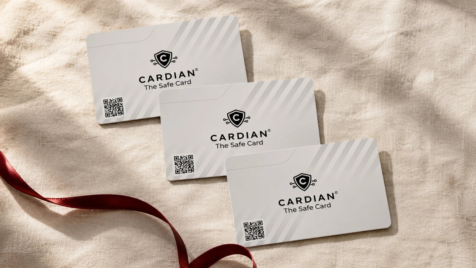 Overhead editorial still life of three identical Cardian cards laid flat on cream linen, slightly fanned with each card offset by about two inches; a single thin oxblood silk ribbon loops loosely around the bottom-left corner of the bottom card and trails off-frame
