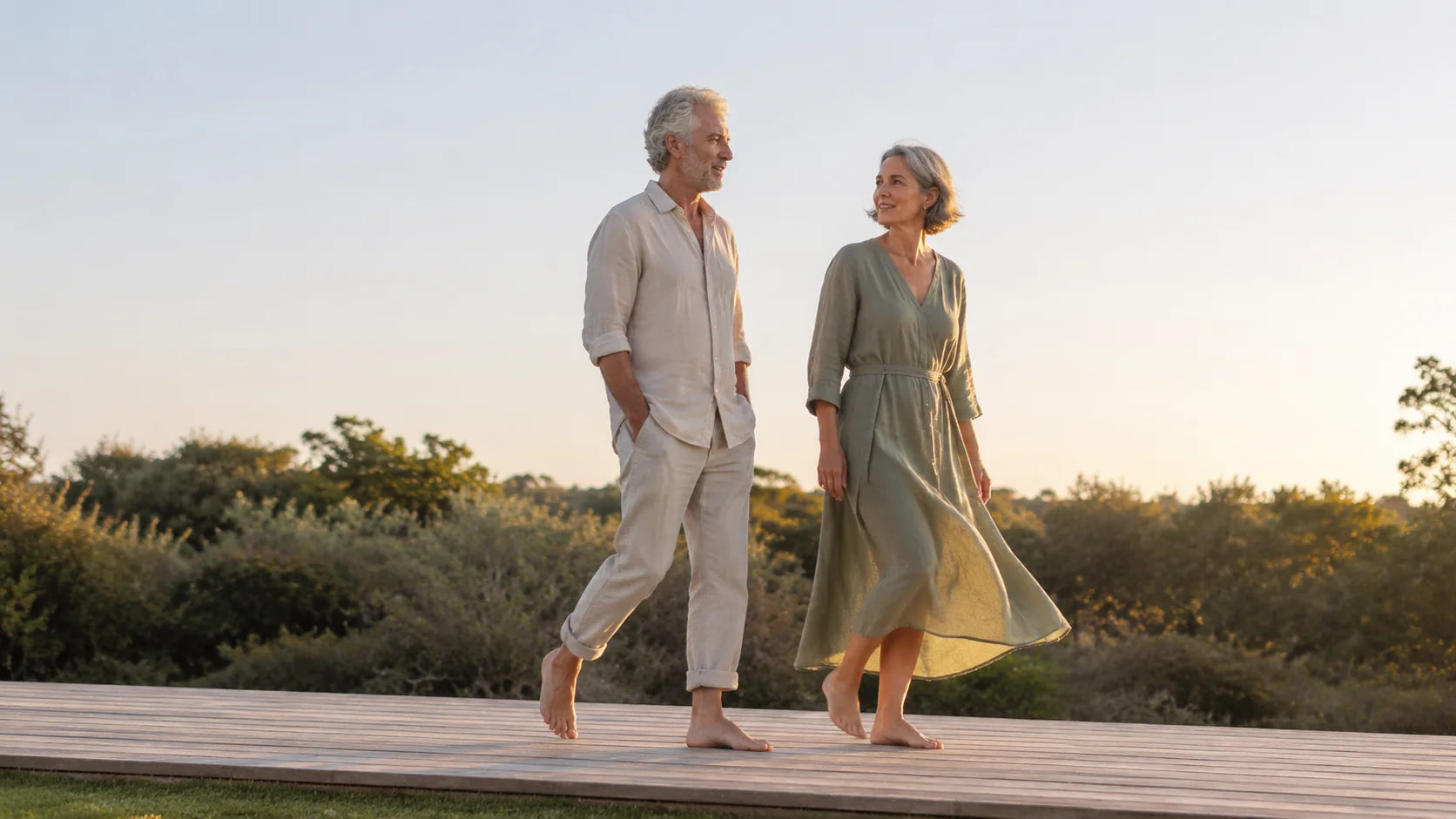 Older man and woman in linen, walking barefoot together along a sunlit wooden deck, candid editorial lifestyle